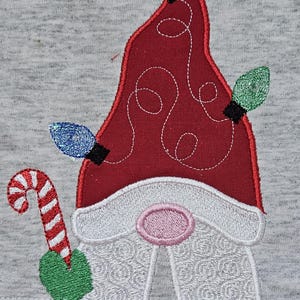 May include: Embroidered gnome with a red hat, white beard, and pink nose. The gnome is holding a candy cane and has a string of lights on its hat.