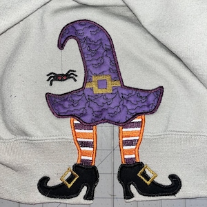 Puede incluir: Sombrero de bruja morado con una araña colgando de él, piernas a rayas naranja y blancas y zapatos negros con hebillas doradas. La bruja está bordada en una sudadera gris claro.