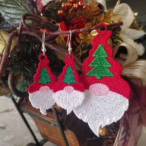 May include: Three pairs of red and white gnome earrings with green Christmas trees on the hats. The earrings are hanging from a branch.