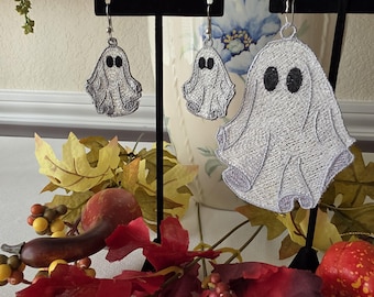 May include: Three silver-toned ghost-shaped earrings featuring black embroidered eyes. The set includes two smaller ghost earrings and one larger ghost earring, all suspended from silver-colored metal hooks on black display stands.