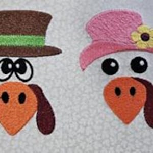 May include: Two felt turkey faces with large eyes and orange beaks. One turkey wears a brown top hat with a green band, the other a pink hat with a yellow flower. The background is a light, speckled pattern.