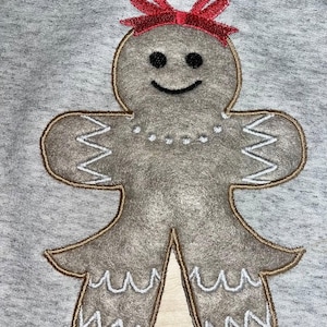 May include: A brown gingerbread person with a red bow on its head. The gingerbread person is embroidered on a gray fabric.