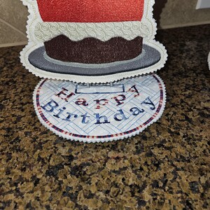 Può includere: Set di sottobicchieri ricamati a forma di torta di compleanno. Il sottobicchiere superiore ha la forma di una torta con glassa rossa e base marrone. Il sottobicchiere inferiore è rotondo con le parole "Happy Birthday" ricamate in blu e rosso.