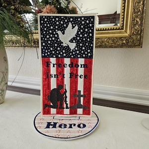 May include: A patriotic tabletop decoration featuring a design reminiscent of the American flag. The top portion has a blue background with white stars, and the bottom has red and white stripes. The text "Freedom isn't Free" is displayed, along with a silhouette of a soldier and a cross. The base reads "Hero".