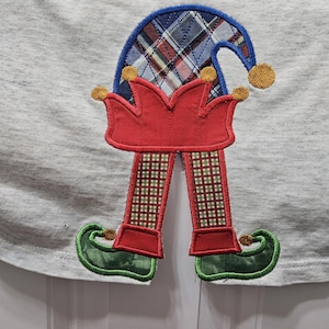 May include: A gray t-shirt with an embroidered elf design. The elf is wearing a red and white plaid hat, red pants, and green shoes.
