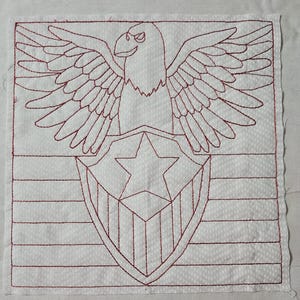 May include: A white square fabric with a red embroidered design. The design features a spread-winged eagle above a shield with a star, and horizontal stripes below. The eagle has a stern expression. The fabric has a dotted pattern.