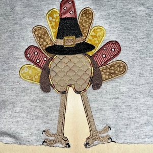 Puede incluir: Diseño de pavo de Acción de Gracias bordado en una tela gris. El pavo lleva un sombrero de peregrino negro con una hebilla marrón y tiene una cola marrón y dorada.