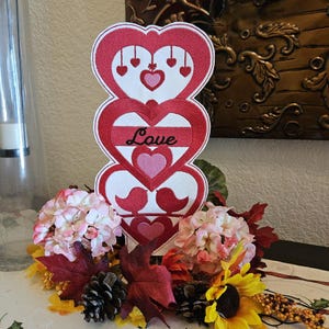 May include: A Valentine's Day decoration featuring three stacked red hearts with white borders. The middle heart has the word "Love" embroidered on it. The decoration is surrounded by pink and red flowers, leaves, and pinecones.