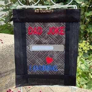 May include: A rectangular fabric garden flag featuring a black border and a dark gray center with a woven pattern. The text DAD JOKE is embroidered in red above a light gray loading bar, a small red heart, and the word LOADING in blue.