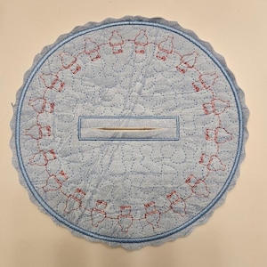 May include: A light blue, circular quilted coaster with a scalloped edge. The coaster features a repeating pattern of red bottle designs around the edge and a rectangular opening in the center. The stitching is light blue.