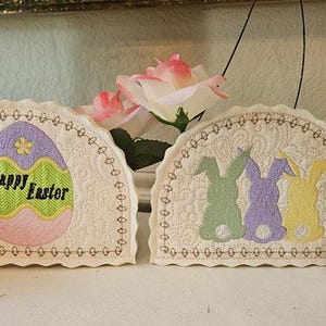 May include: Embroidered Easter decorations featuring an egg with "Happy Easter" text and three bunny silhouettes. The decorations are cream-colored with pastel accents, and are displayed with decorative Easter eggs and flowers.