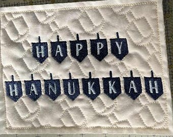 May include: A white quilted fabric square with the words "HAPPY HANUKKAH" embroidered in navy blue, each letter within a dreidel shape. The fabric has a stitched, repeating pattern.