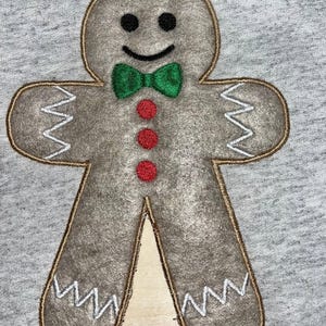 May include: A grey gingerbread man applique with a brown outline, black eyes, a green bow tie, and three red buttons. The gingerbread man has white zig-zag stitching on the arms and legs. The applique is on a grey background.