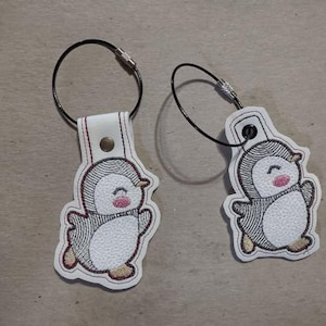 Può includere: Due portachiavi ricamati a forma di pinguino. Ogni portachiavi presenta un pinguino bianco con dettagli neri e grigi, un rossore rosa e un anello di filo nero. Il pinguino è attaccato a un'etichetta di pelle bianca con una chiusura in metallo.