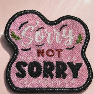 Può includere: Toppa ricamata rosa con le parole "Sorry Not Sorry" in bianco e nero. La toppa ha un bordo nero e un aspetto strutturato. Piccoli accenti di foglie verdi sono sui lati della parola "Sorry".