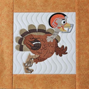 May include: Embroidered turkey wearing a football helmet, holding a football, and running. The turkey is brown, with a brown and tan tail. The helmet is orange and brown. The background is white with a wavy pattern, framed by orange fabric.