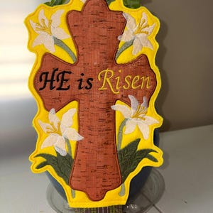 May include: Embroidered Easter cross with the text "HE is Risen". The cross is brown with a yellow border and white lily flowers. The design is on a yellow background.
