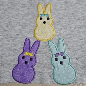 Può includere: Tre forme di coniglio in feltro ricamate su uno sfondo di tessuto grigio. I conigli sono gialli con pois bianchi, viola con motivi a spirale e blu con pois bianchi. Ogni coniglio ha un naso nero, occhi neri e un fiocco blu.