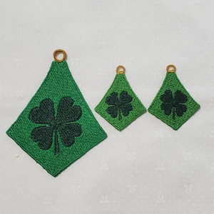Op de afbeelding: Drie groene vilt shamrock ornamenten met gouden rand. De shamrocks zijn van verschillende grootte en hebben de vorm van een ruit.