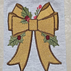 May include: A gold embroidered bow with green holly leaves and red berries. The bow is on a light gray background.