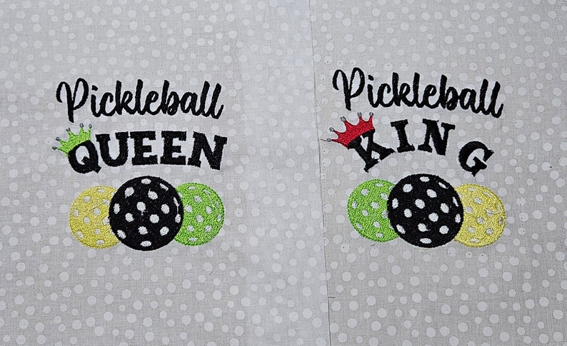 May include: Two embroidered towels with the words "Pickleball Queen" and "Pickleball King" with a crown above each word. Each towel has three embroidered pickleball paddles.
