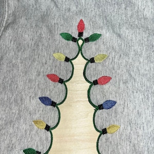 Puede incluir: Una sudadera gris con un diseño de árbol de Navidad bordado. El árbol está hecho de luces de cadena verdes con bombillas rojas, amarillas, azules y verdes.