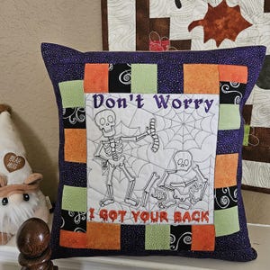 May include: Square Halloween-themed pillow with a purple background and a patchwork border of orange, green, and black squares. The center features a white panel with a skeleton design and the text "Don't Worry I Got Your Back" embroidered in purple and orange.