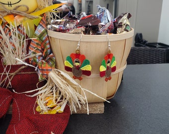 May include: A pair of turkey-shaped earrings with green, yellow, red, and brown embroidery. The earrings are hanging from a wooden basket filled with chocolate candy bars.