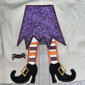 May include: Embroidered patch of a witch's legs with black shoes, purple and orange striped stockings, and a purple skirt with a bat pattern. A small black spider with red eyes hangs from a string.