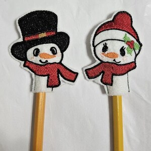 Op de afbeelding: Twee potloodtoppers in de vorm van sneeuwmannen. Eén sneeuwman draagt een zwarte hoge hoed met een rode band en een rode sjaal. De andere sneeuwman draagt een rode muts met hulst en een rode sjaal. Beide hebben oranje neuzen en zwarte ogen.