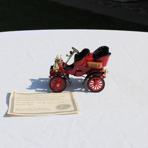 May include: A miniature red vintage-style automobile model featuring black seats, gold-toned accents, and red spoked wheels. A small tan suitcase is attached to the side. Beside the model sits a rectangular certificate of authenticity document.