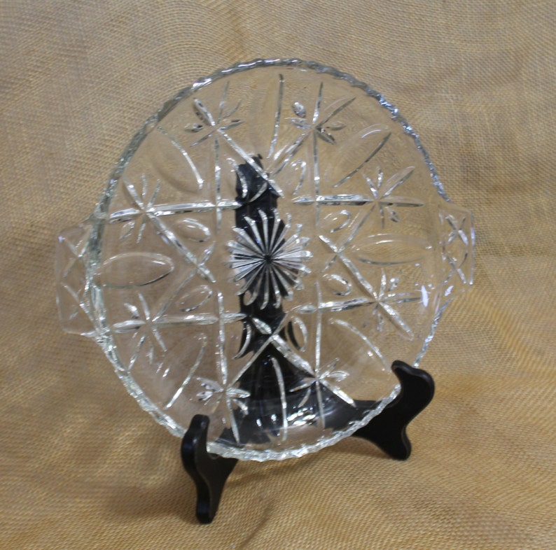 May include: A clear glass serving dish with a starburst pattern and two handles. The dish is on a black stand.