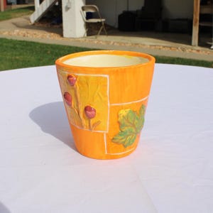 May include: An orange ceramic flower pot with a cream-colored interior. The pot features decorative squares with red flower and green leaf designs. The pot is sitting on a white surface.