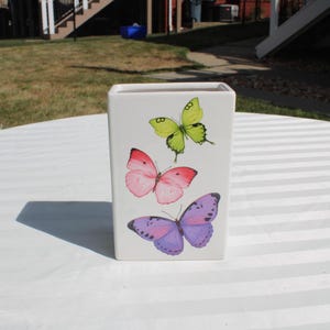 May include: A white rectangular vase decorated with three colorful butterflies. The butterflies are in shades of green, pink, and purple. The vase is sitting on a white striped surface.