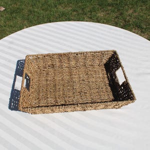 May include: A rectangular woven basket tray with built-in handles. The tray is made of natural-colored woven material and is set on a white and gray striped surface. The basket is empty.