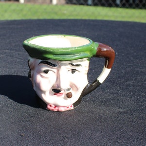 May include: A ceramic mug shaped like a man's face with a green hat, brown mustache, and a pipe. The mug has a brown handle.
