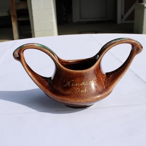 May include: A brown ceramic pitcher with two arched handles. The pitcher has a dark interior and a lighter brown exterior with a glossy finish. The words "Windsor Ont." are inscribed on the front in gold lettering.