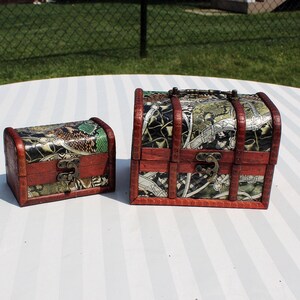 May include: Two decorative wooden treasure chests. The chests have a dark brown frame with a patterned top and sides. The smaller chest is on the left, and the larger chest is on the right. Both chests have a metal clasp.