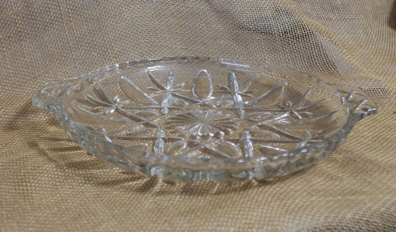 May include: A clear glass serving dish with a scalloped edge and a starburst pattern. The dish has two handles on the sides.