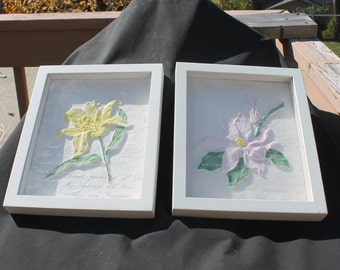 May include: Two framed floral art pieces. One features a yellow lily, the other a pink flower, both with green leaves. The flowers are set against a textured white background within white wooden frames. Decorative wall art.