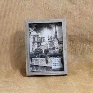 May include: A rectangular picture frame with a weathered gray finish containing a black and white photograph of the Notre-Dame Cathedral in Paris. The image features the cathedral facade, a stone wall, and a river, with a small white label in the corner.