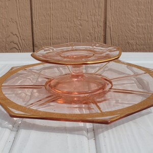 May include: A two-tiered glass serving dish with a pink hue and gold trim. The top tier is smaller and sits on a pedestal. The bottom tier is larger and has a scalloped edge.
