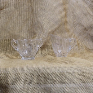 May include: A clear glass sugar bowl with two handles and a matching creamer. Both pieces have a floral pattern and are set against a neutral background. The sugar bowl is approximately 3 inches wide.