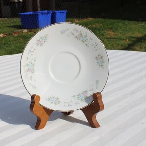 May include: A white saucer with a gold rim and a floral design featuring pink and blue flowers. The saucer is on a wooden stand.