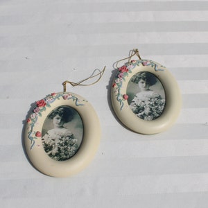 May include: Two oval-shaped, cream-colored picture frames with a floral design. Each frame has a small, black and white photo of a young girl wearing a white dress. The frames have a gold string for hanging.