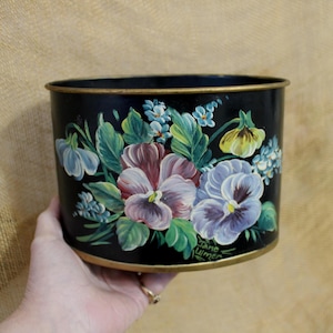 May include: Black metal planter with a hand-painted floral design. The planter features pansies in shades of purple, white, and yellow, with green leaves. The rim and base have a gold trim. The artist's signature is visible.