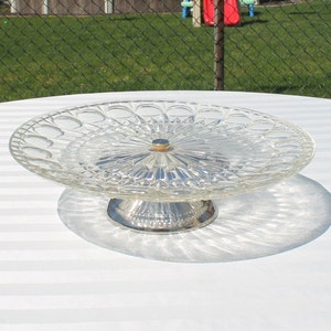 May include: A clear glass cake stand with a silver-colored base. The stand features a decorative, patterned glass plate with a scalloped edge. The base is round and has a detailed, textured design. The stand is on a white striped surface.