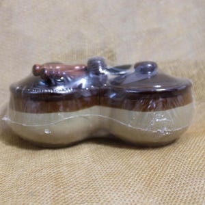 May include: A ceramic double-dish container with a brown and tan color scheme, wrapped in clear plastic. The container has two rounded compartments, each with a lid. The lids and the top portion of the container are brown, while the bottom is tan.