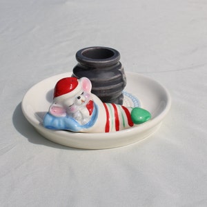 May include: A white ceramic dish with a sleeping mouse figurine wearing a Santa hat and striped blanket. A small, dark gray vase sits on the dish. The mouse is lying on a blue pillow with a green foot. A festive, holiday-themed decorative item.