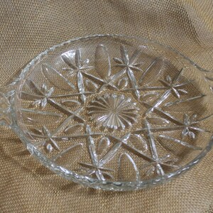May include: A clear glass serving dish with a starburst pattern and two handles. The dish is on a brown burlap background.